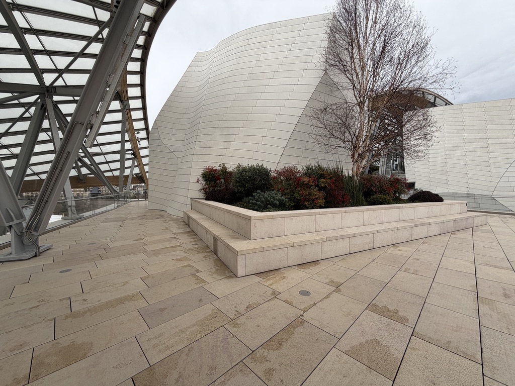 Terrasse pavée avec marches et jardinière au pied d’une façade courbe de la Fondation Louis Vuitton.