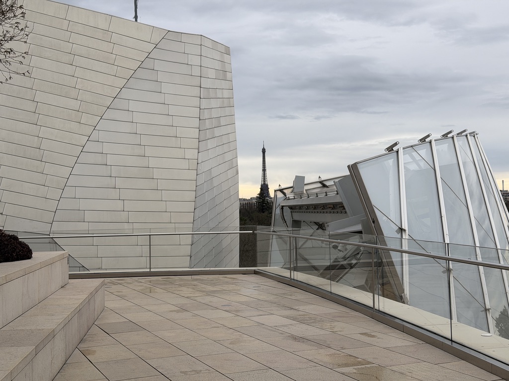 Vue depuis une terrasse de la Fondation Louis Vuitton, entre une façade courbe et les voiles de verre, avec la tour Eiffel au loin.